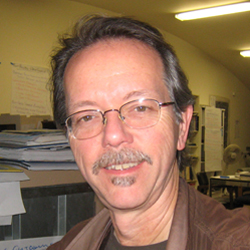 Brian Beveridge smiles, wearing wire-framed glasses and a brown jacket.