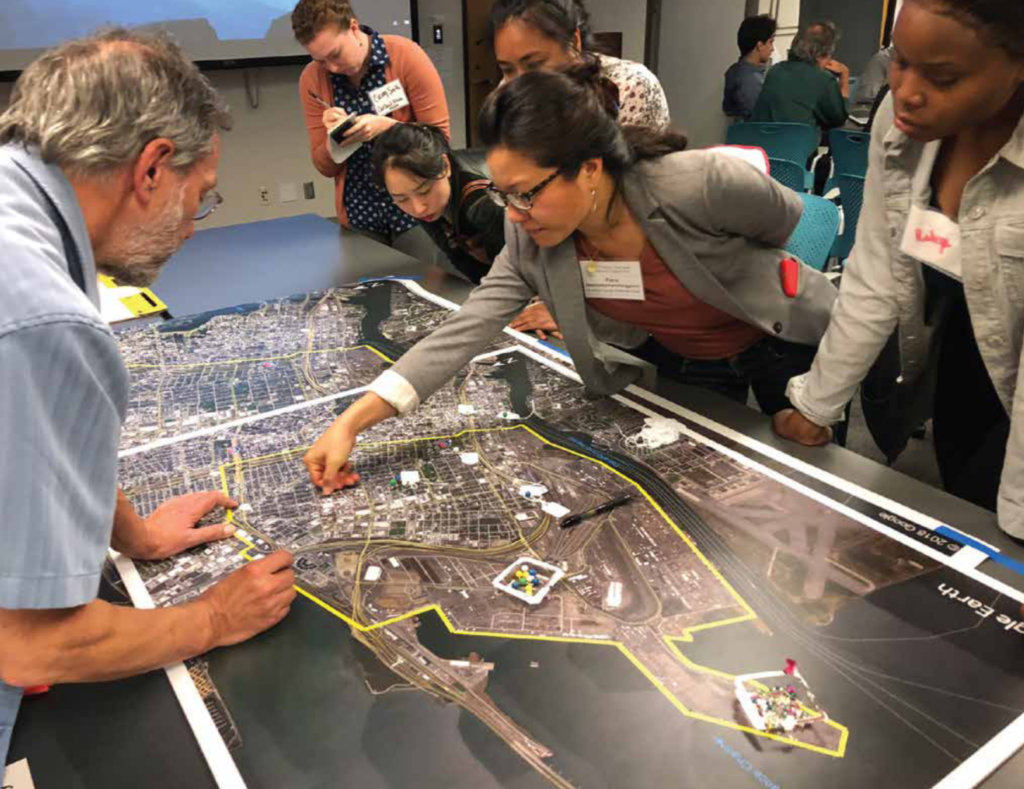 West Okland residents and air quality experts stand around a large table-top map, while a woman with dark hair and glasses points to the center of it.