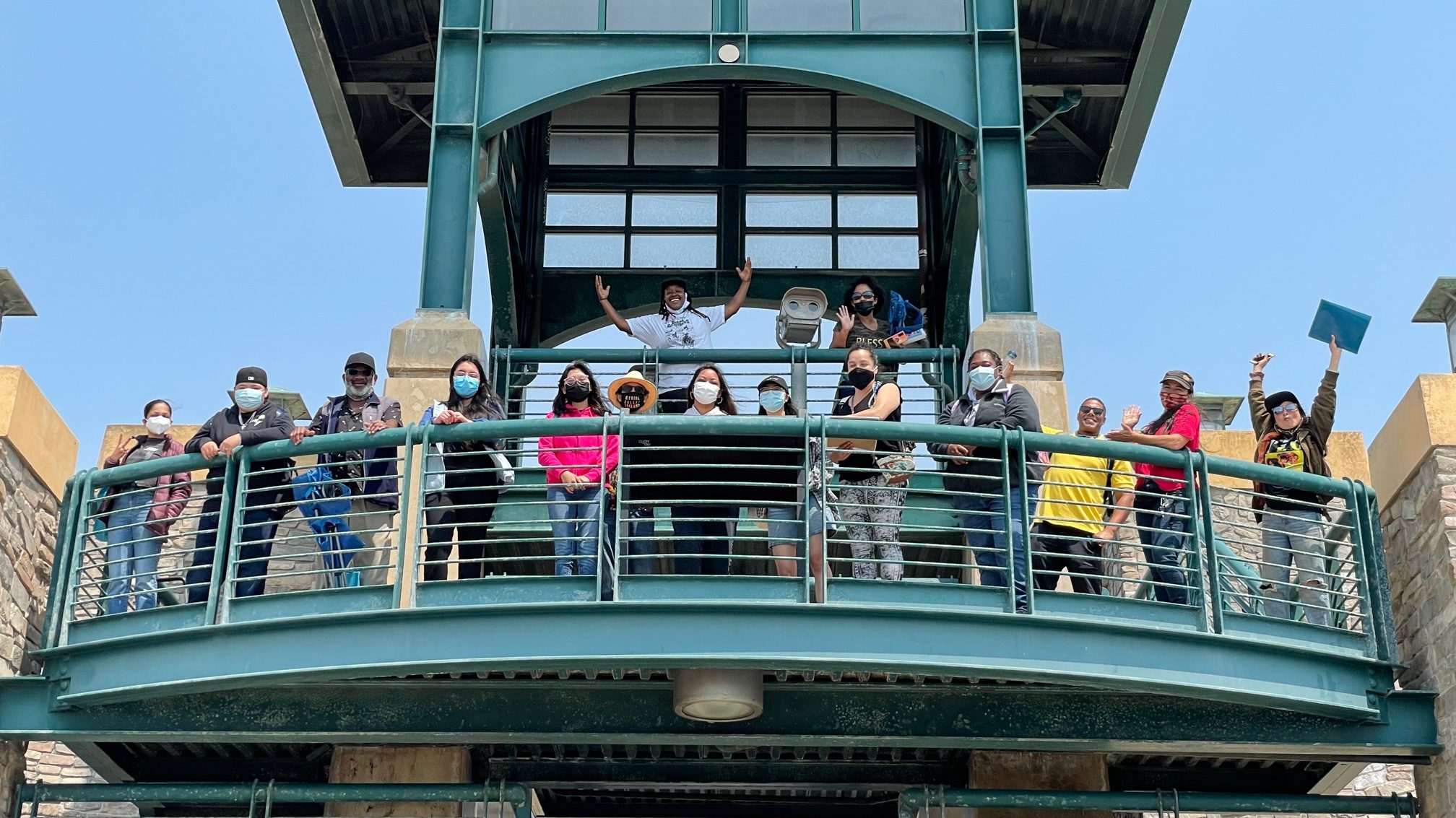 15 students of mixed ages stand atop a large green viewing deck behind a green metal railing, smiling and waving at the camera on a sunny afternoon.