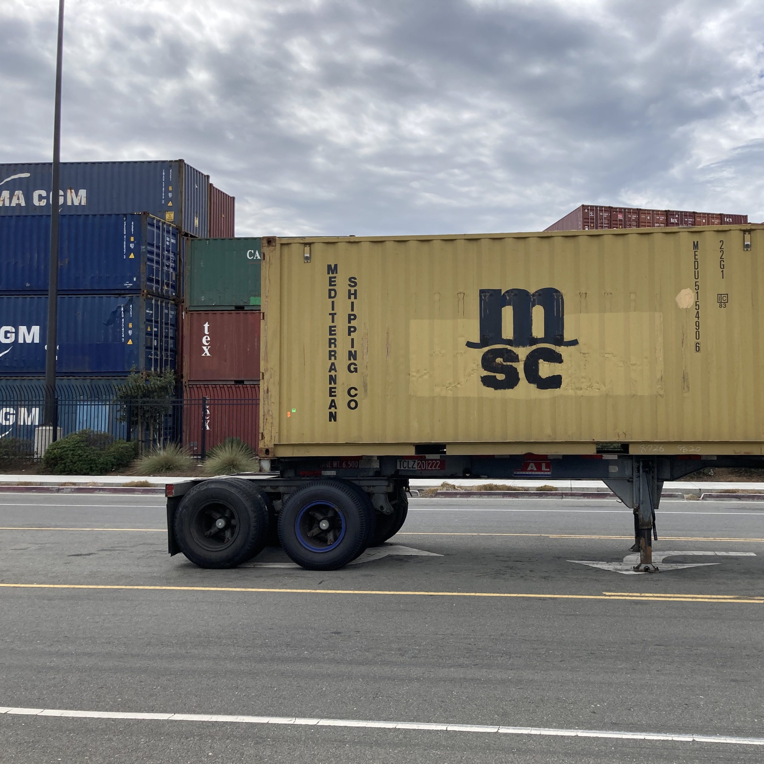 A yellow commercial truck trailer sits unattached in front of a wall of stacked shipping containers. 
