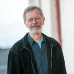 Brian stands outside, smiling, in a black jacket and blue collared shirt.