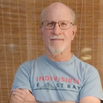 Aaron wears glasses and a gray t-shirt as he gazes into the camera with arms crossed.