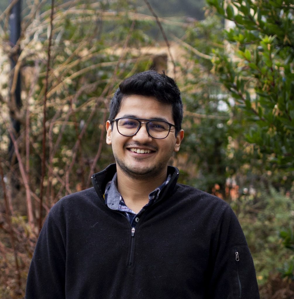 Meet smiles in front of a backdrop of trees, wearing dark-rimmed glasses and a dark pullover fleece jacket.