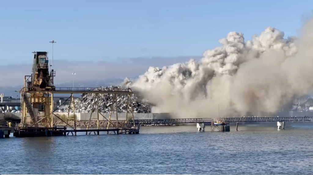 A massive cloud of thick gray smoke bellows from a huge pile of scrap metal waste along the Oakland waterfront.
