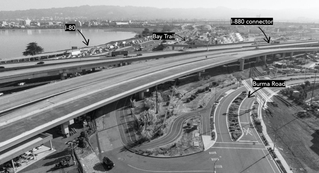 Black-and-white aerial photo showing the I-880 freeway ramp connecting to I-80 with the rest of Oakland and Emeryville in the far distance. Burma Road sweeps along the underside of the freeway in a curved loop towards the water.