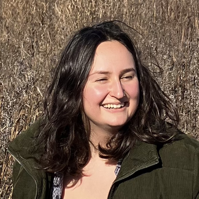 Clara stands in front of a marsh on a sunny day, smiling wide in a dark green jacket and long brown hair.