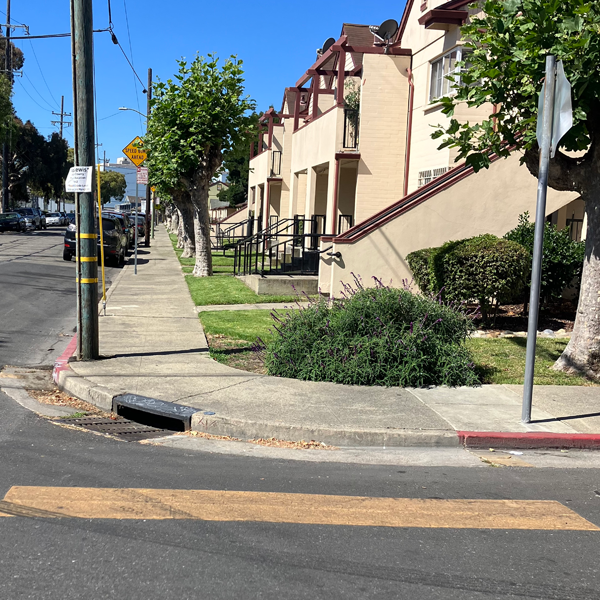 The view from a street intersection looking at the corner of a row of townhouses to the right, flanked by a sidewalk. There is a sewer drain at the sidewalk's corner, but no sidewalk ramps cut into the curb for pedestrians.