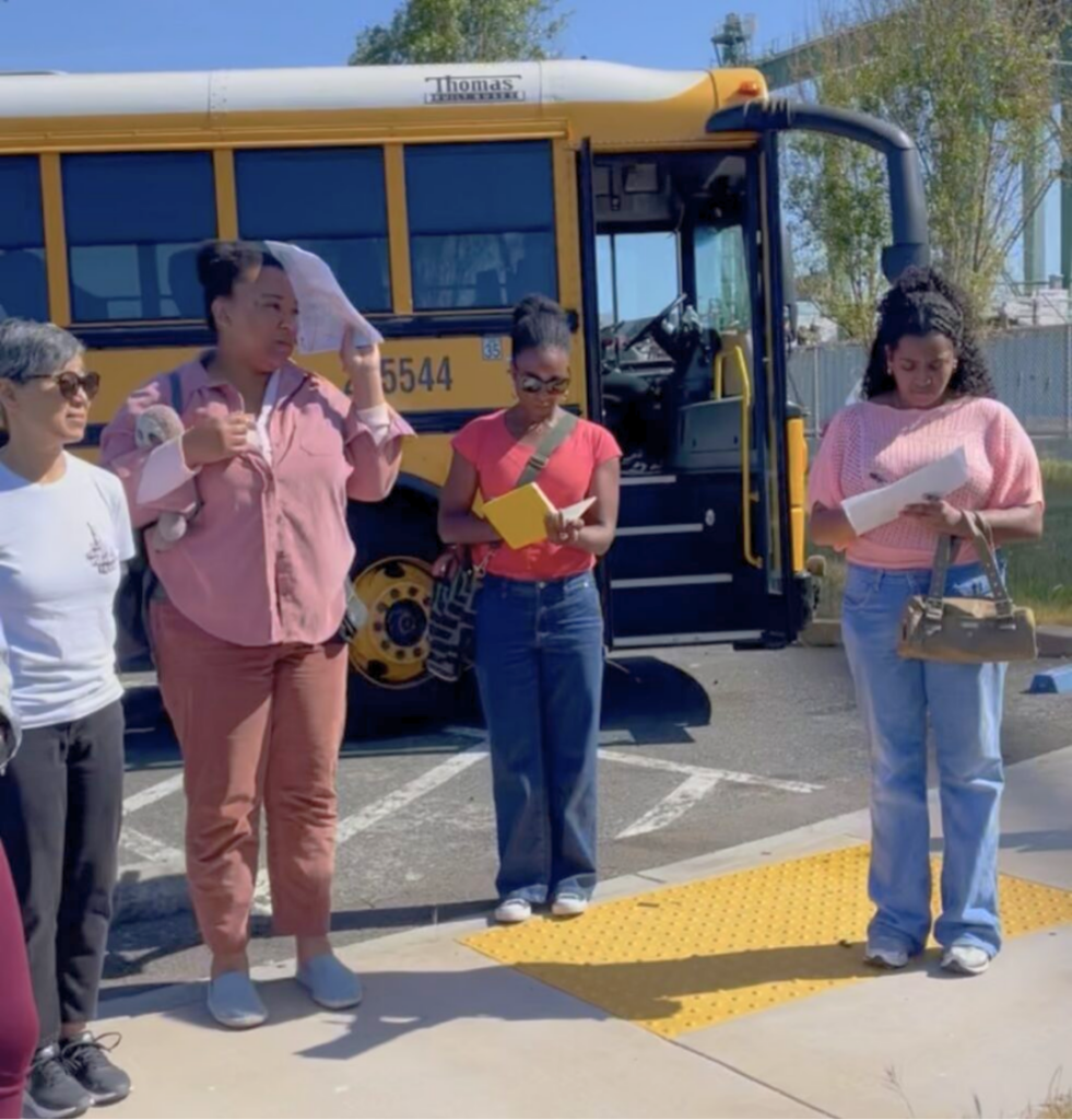 Four people stand in front of a school bus. Two people take notes.